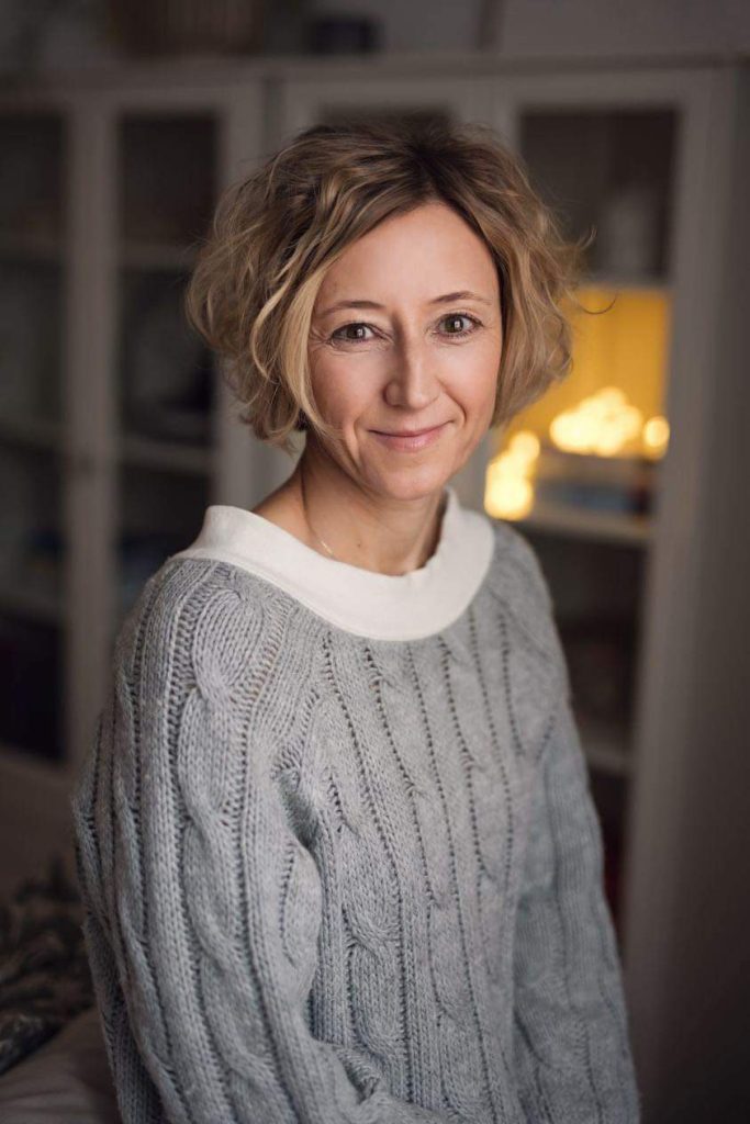 Portret kobiety z krótkimi, kręconymi blond włosami, ubranej w szary, gruby sweter o splotach warkoczowych. Kobieta uśmiecha się delikatnie, a w tle widać rozmyte wnętrze z delikatnym oświetleniem.